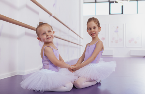 Two young girls wearing purple tutus sit on the floor of a dance studio