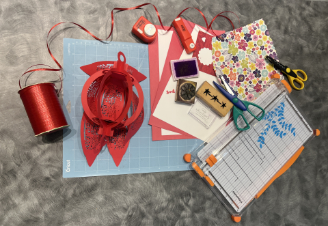 A collection of paper crafting tools including a Cricut mat, red ribbon, a paper cutter, red and white construction paper and stamps.
