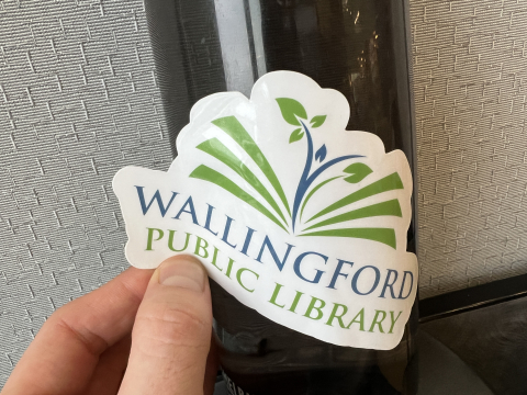 A green and blue Wallingford Public Library logo sticker being placed onto a water bottle by a hand.