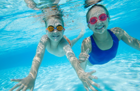 two children swimming underwater, wearing goggles, and smiling at camera