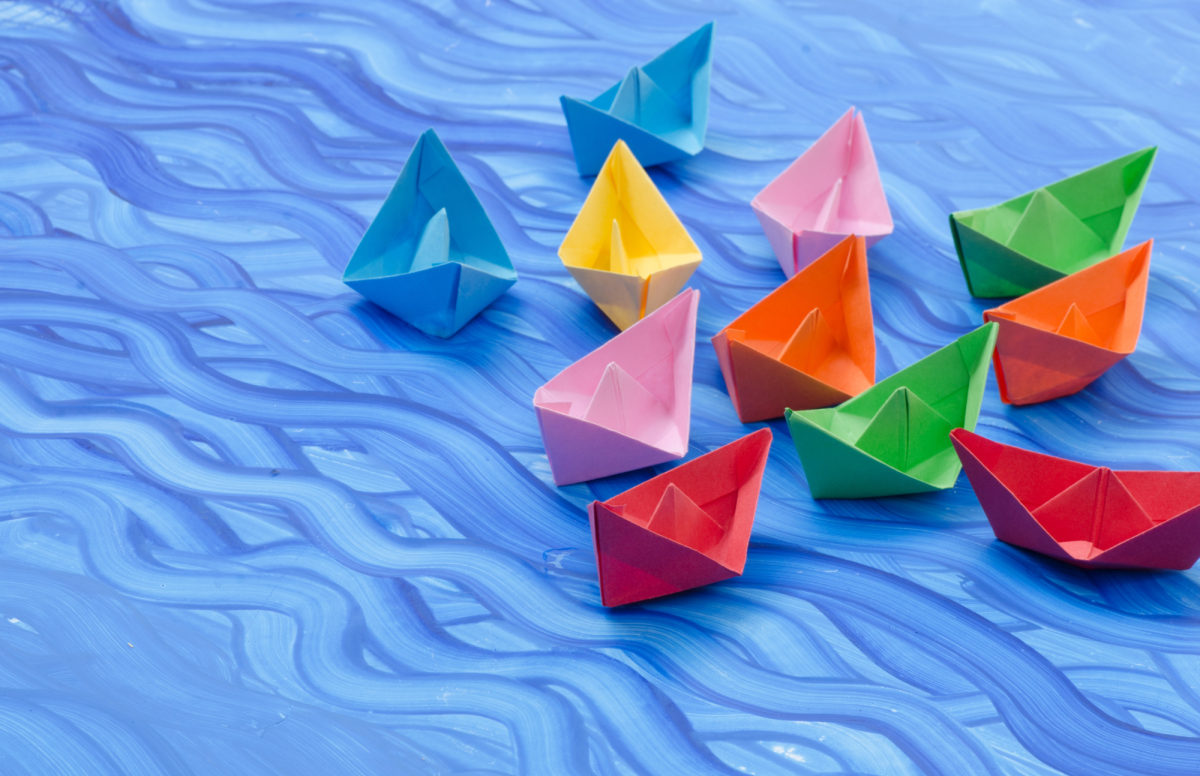 11 Paper folded boats sitting on a painted piece of paper that has waves like the sea.